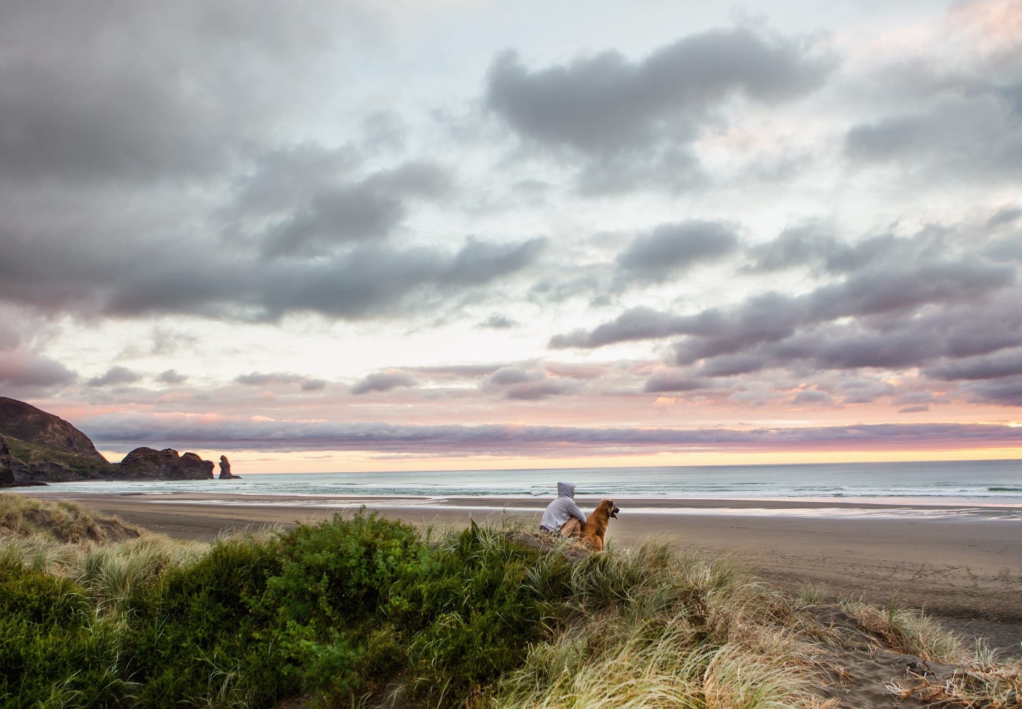 Man in a hoodie sitting alone with his dog at the beach watching the sunset.