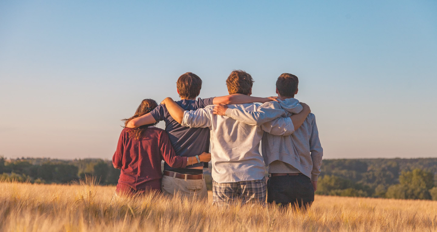 Three young men and a young woman holding each other by the shoulders in a wheat field under the blue sky back facing the camera.